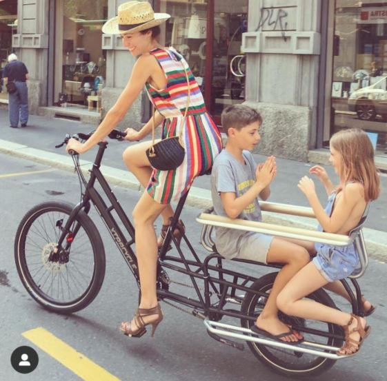 Woman biking in heels with two children facing each other and playing "patty cake" on the back of the bike